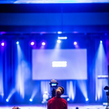 Cameraman capturing a live event on stage with colorful lights and blurred audience.
